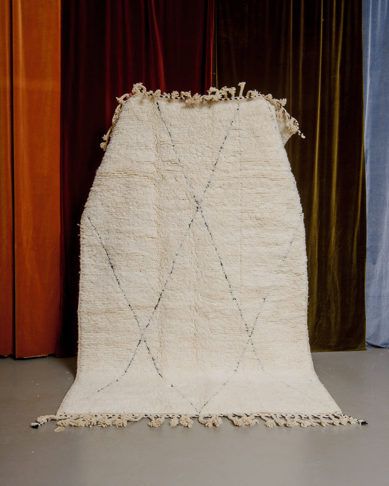 Berberlin Beniourain rug in cream and black wool being held up in front of velvet curtains.