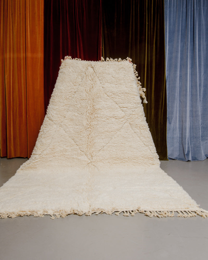 Berberlin Beniourain rug in cream wool being held up in front of velvet curtains.
