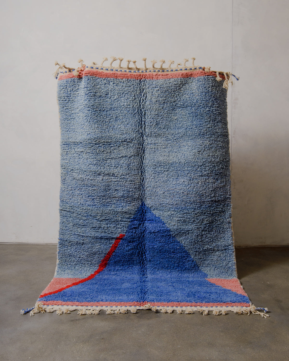 Modern rug in shades of blue, pink and red being held up in front of a white wall. 