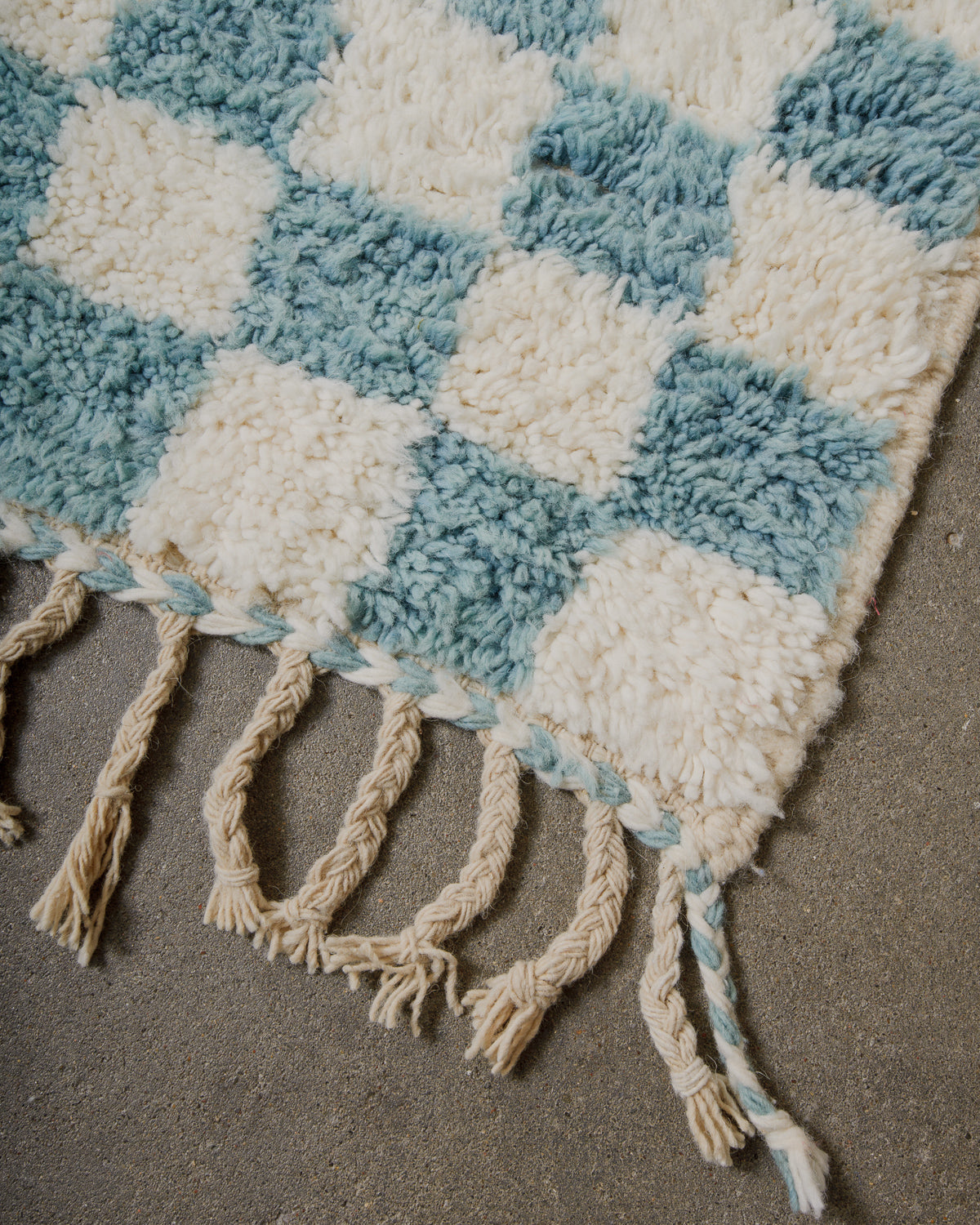 Corner of checkered wool rug in cream and robin's egg blue, with natural wool tassels on concrete floor. 