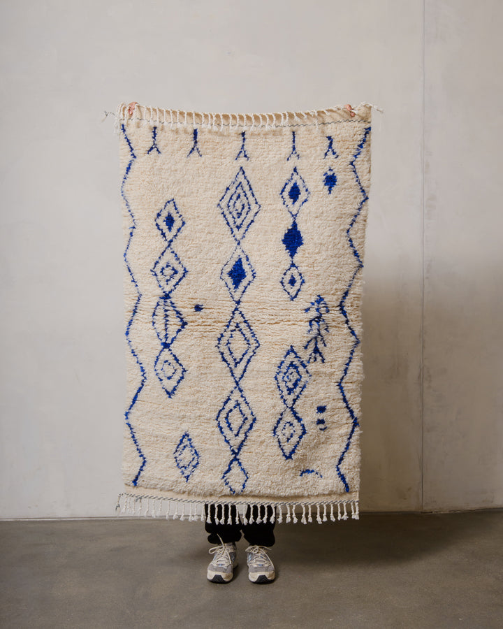Berberlin Azilal rug in cream wool with blue cotton  being held up in front of a white wall.