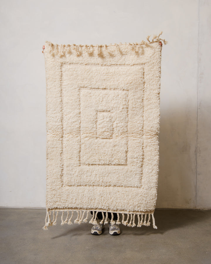 Small Berberlin Beniourain rug in cream wool with geometric woven design, being held up in front of a white wall. 