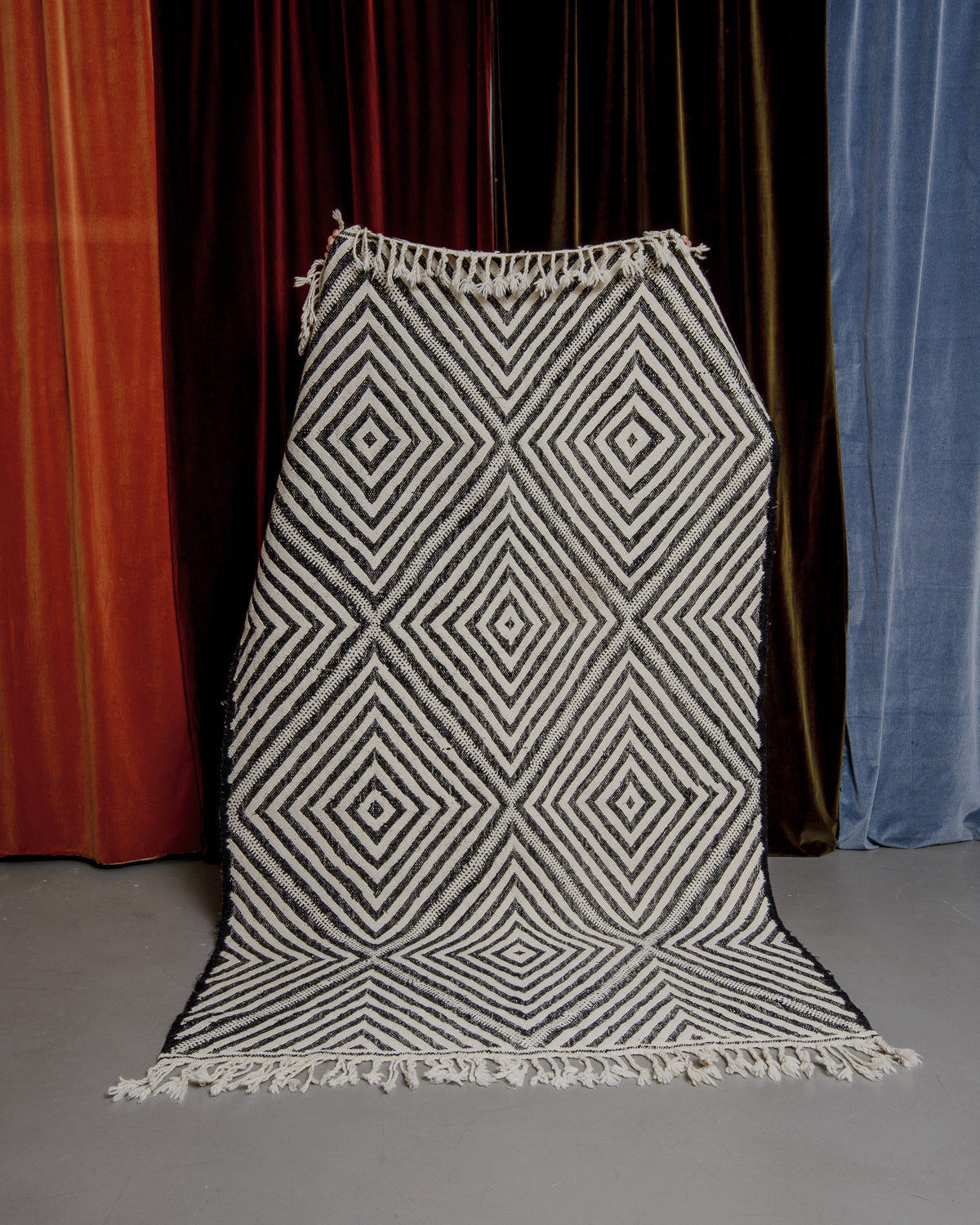 Black and white kelim rug with diamond pattern being held up in front of velvet curtains.