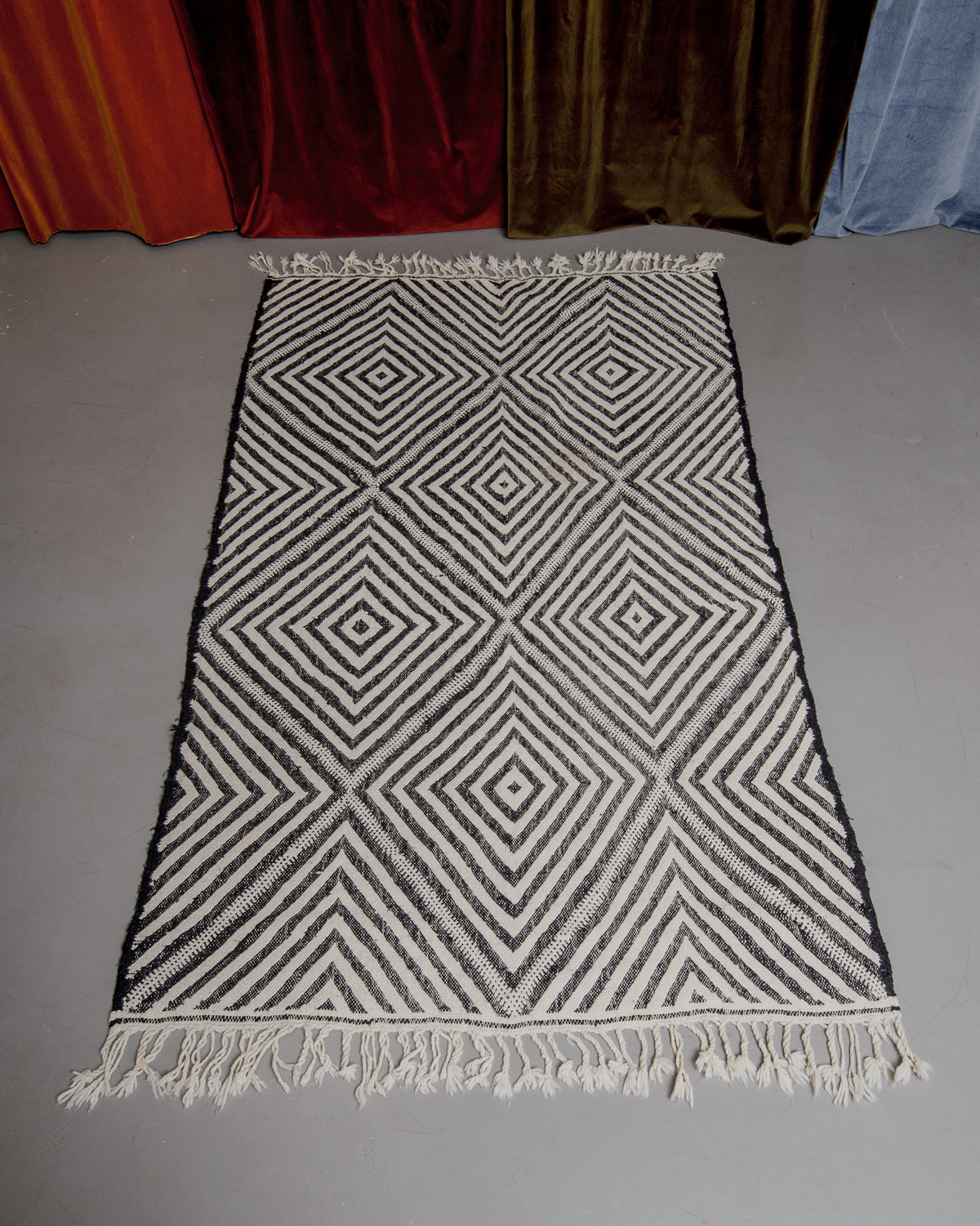 Black and white kelim rug with diamond pattern laying on floor in front of velvet curtains.