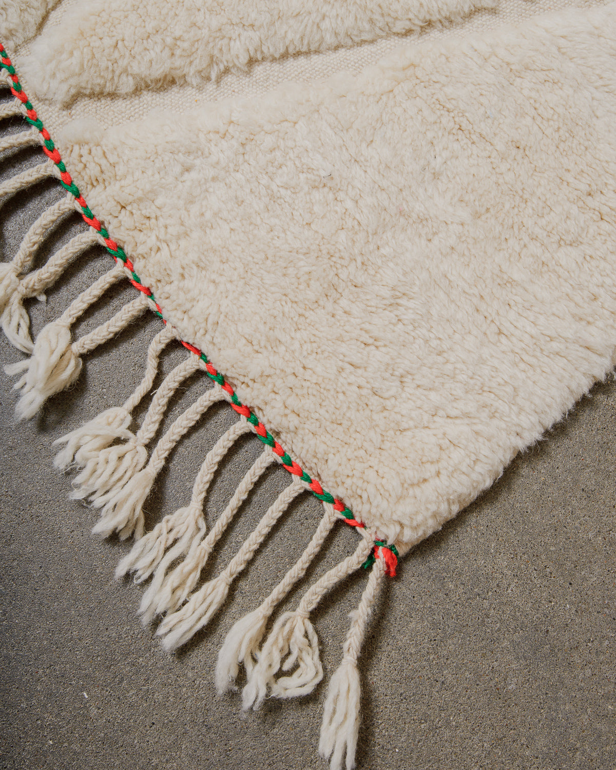 Corner of cream wool rug with red and green trim and natural wool tassels. 