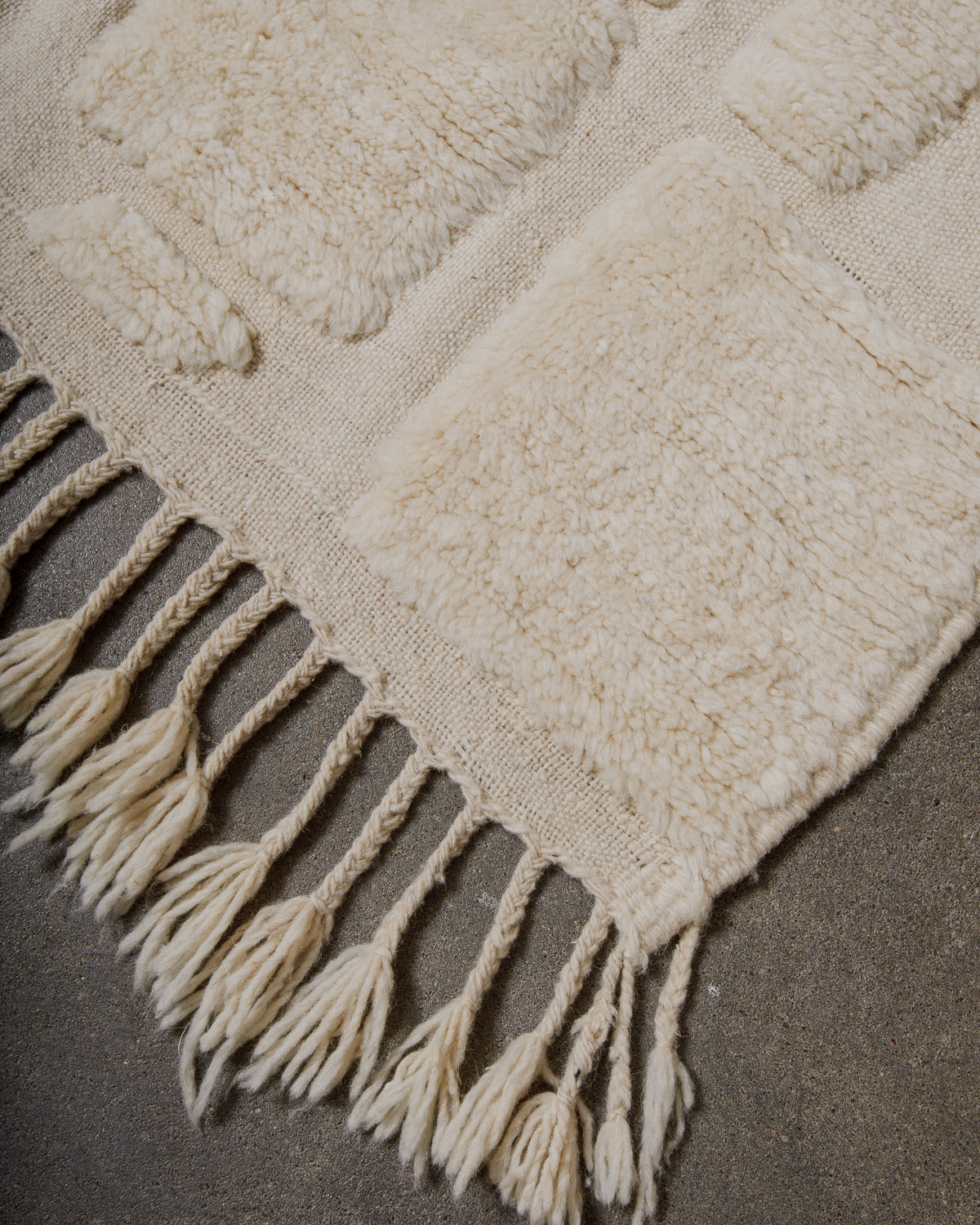Close-up of a textured beige wool rug with tassels on a cement surface .