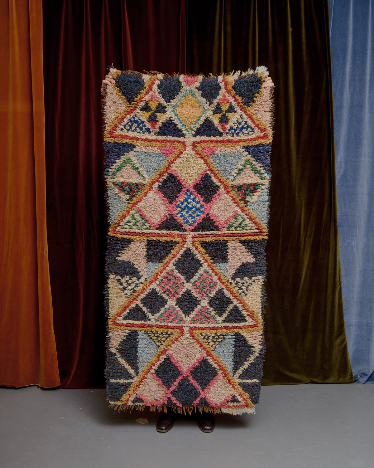 Vintage Berberlin boucherouite runner rug being held up in front of a velvet curtain. 