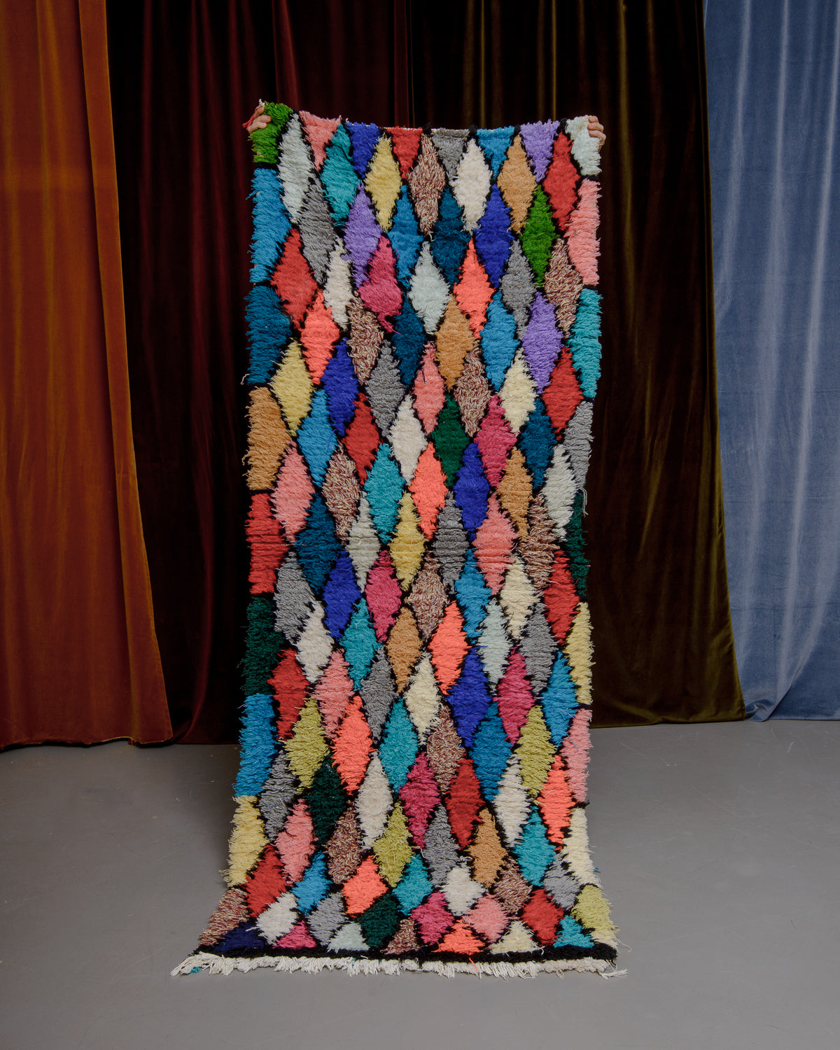 Vintage Berberlin boucherouite runner rug being held up in front of a velvet curtain. 