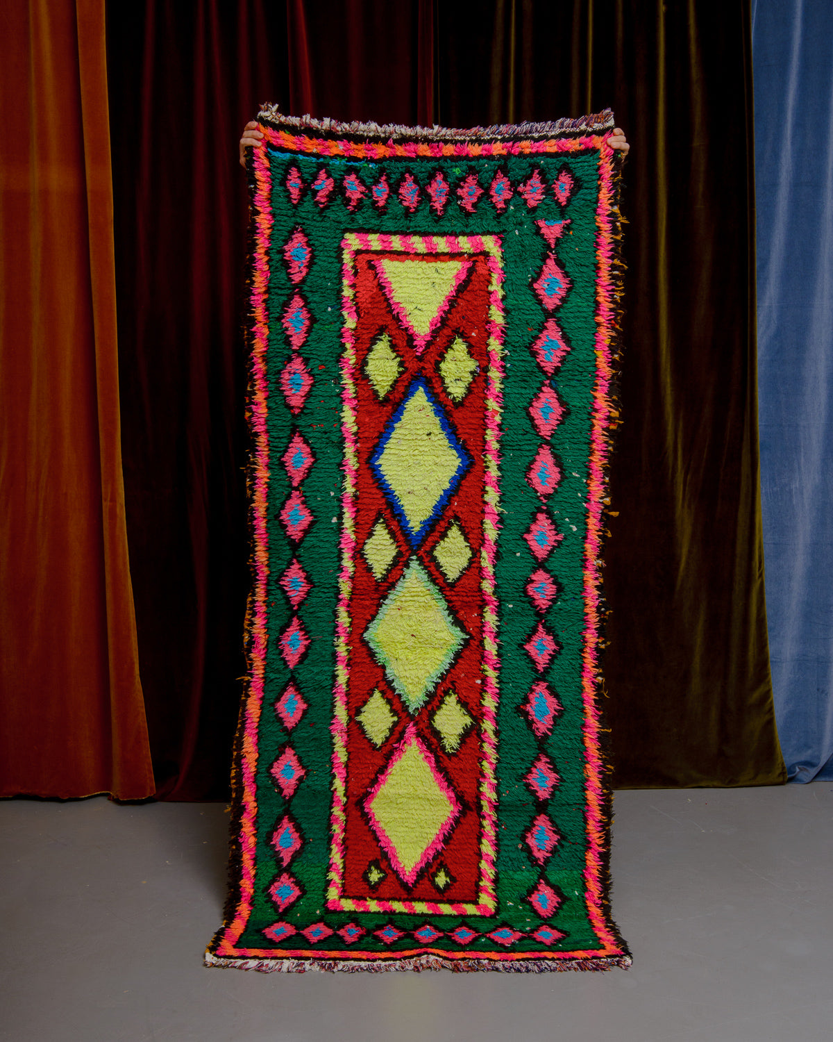 Vintage Berberlin boucherouite runner rug being held up in front of a velvet curtain. 