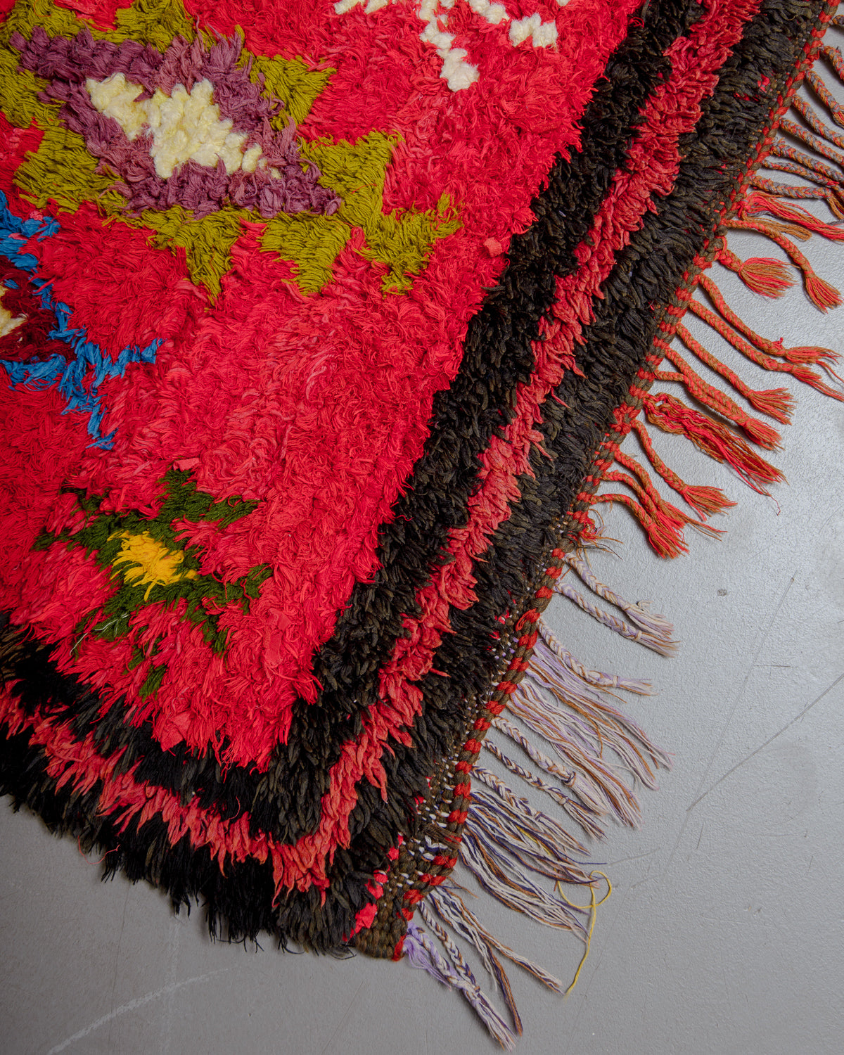Corner of red , black, green and yellow rug with multicoloured cotton tassels.