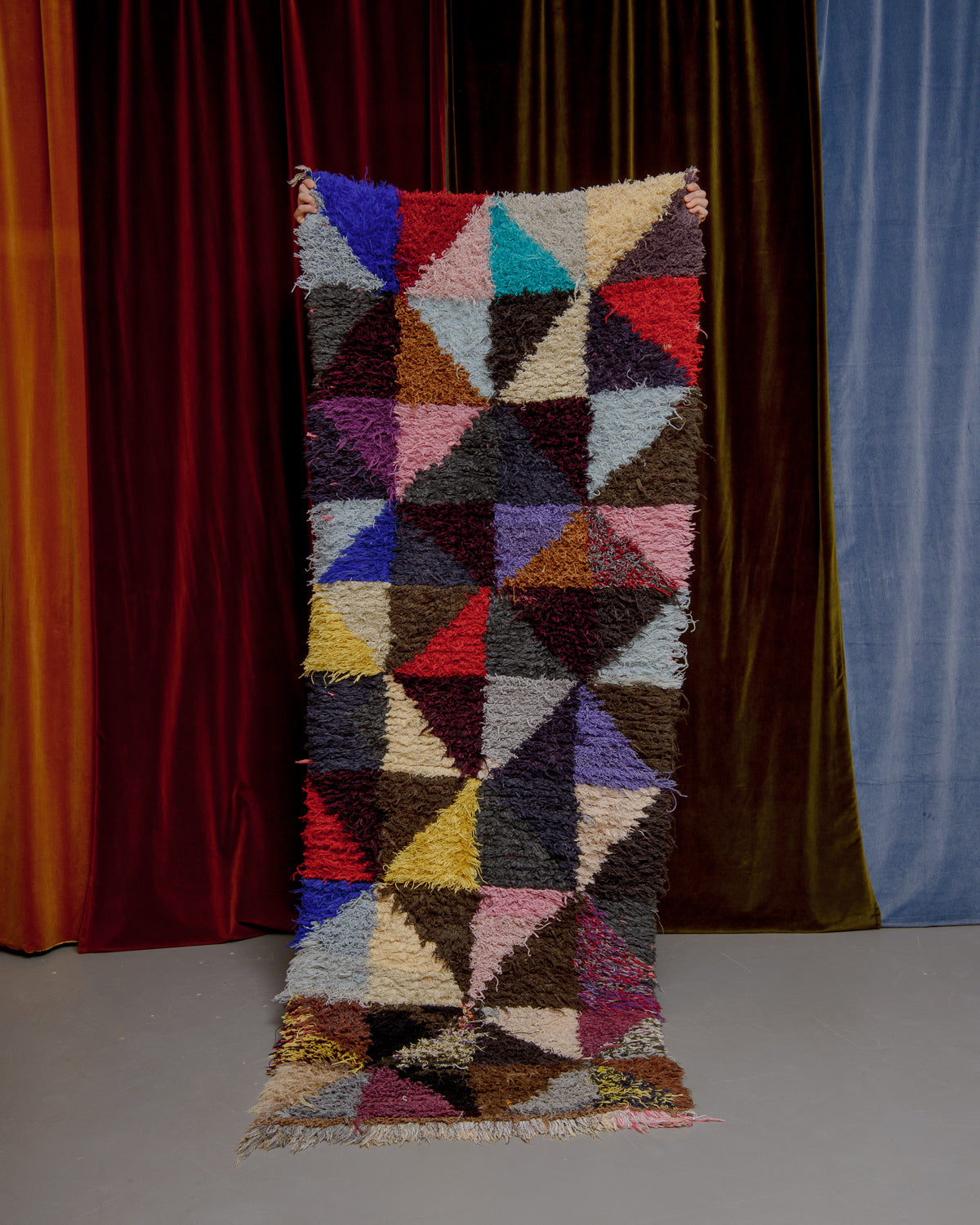 Vintage Berberlin boucherouite runner rug being held up in front of a velvet curtain. 