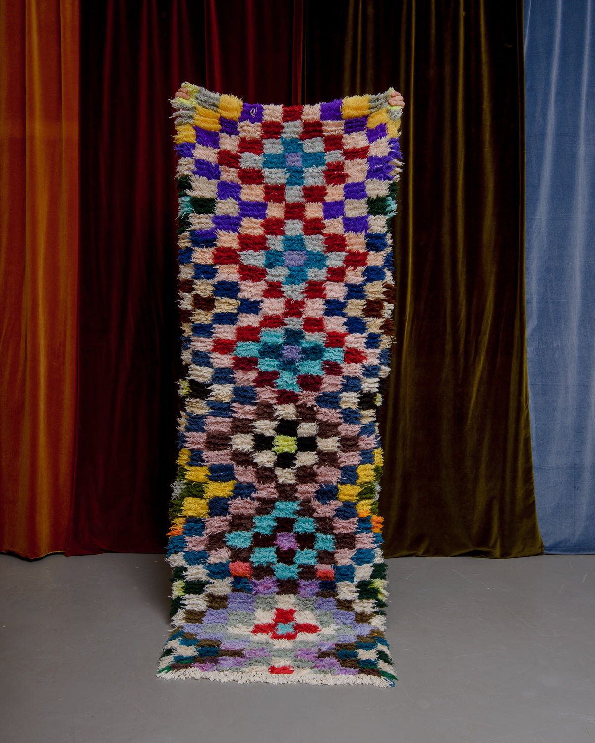 Vintage Berberlin boucherouite runner rug being held up in front of a velvet curtain. 