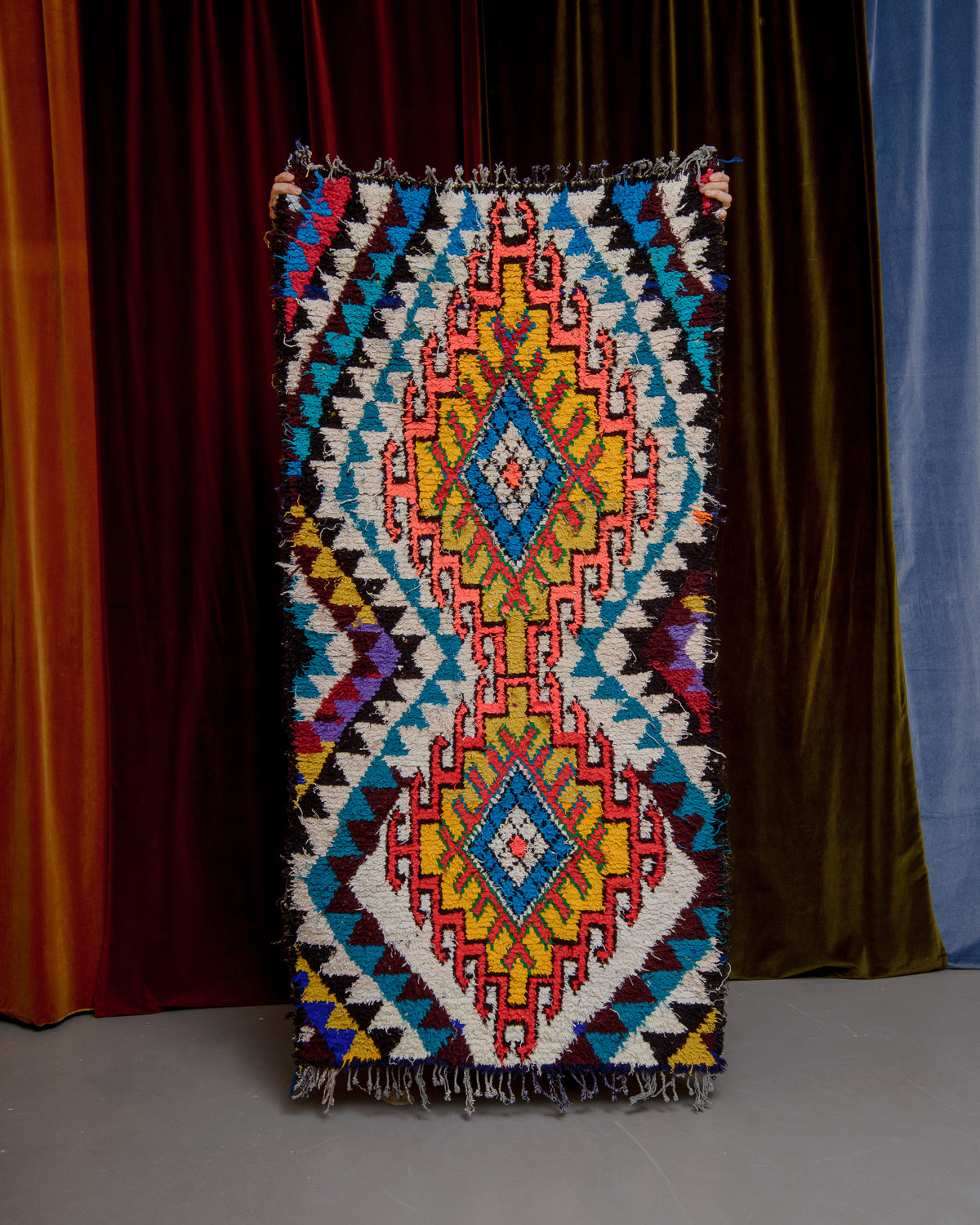 Vintage Berberlin boucherouite runner rug being held up in front of a velvet curtain. 