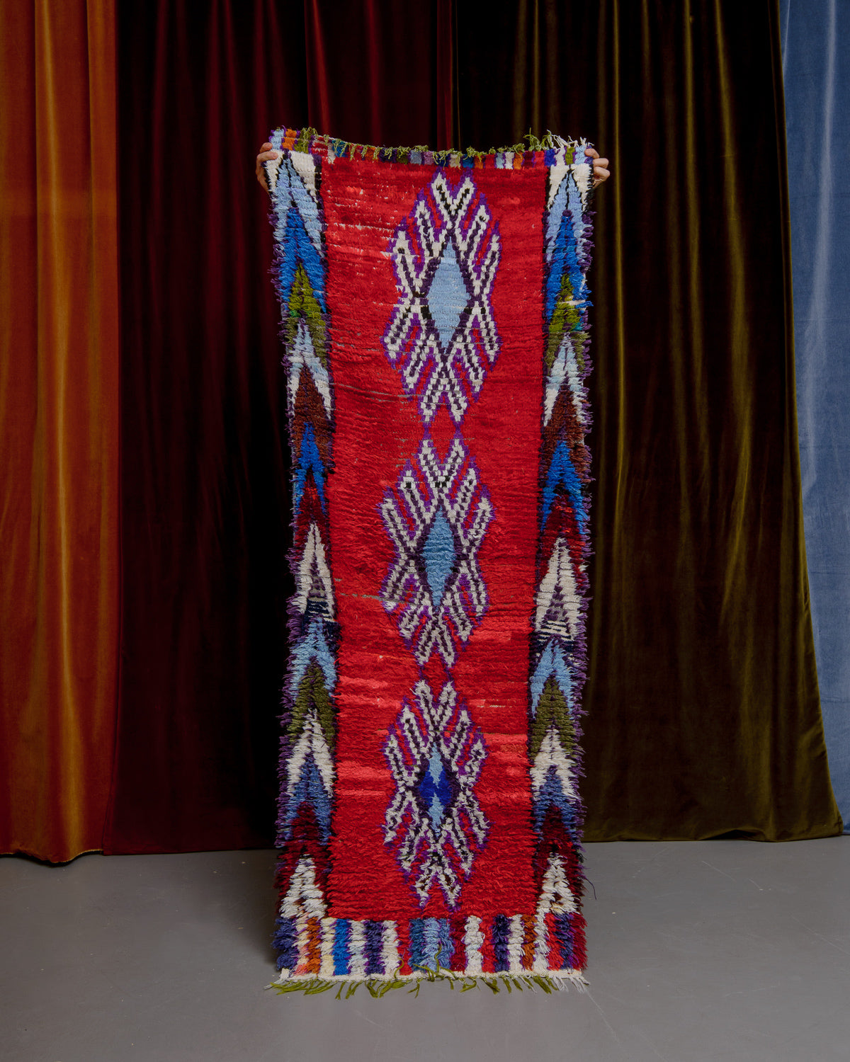 Vintage Berberlin boucherouite runner rug being held up in front of a velvet curtain. 