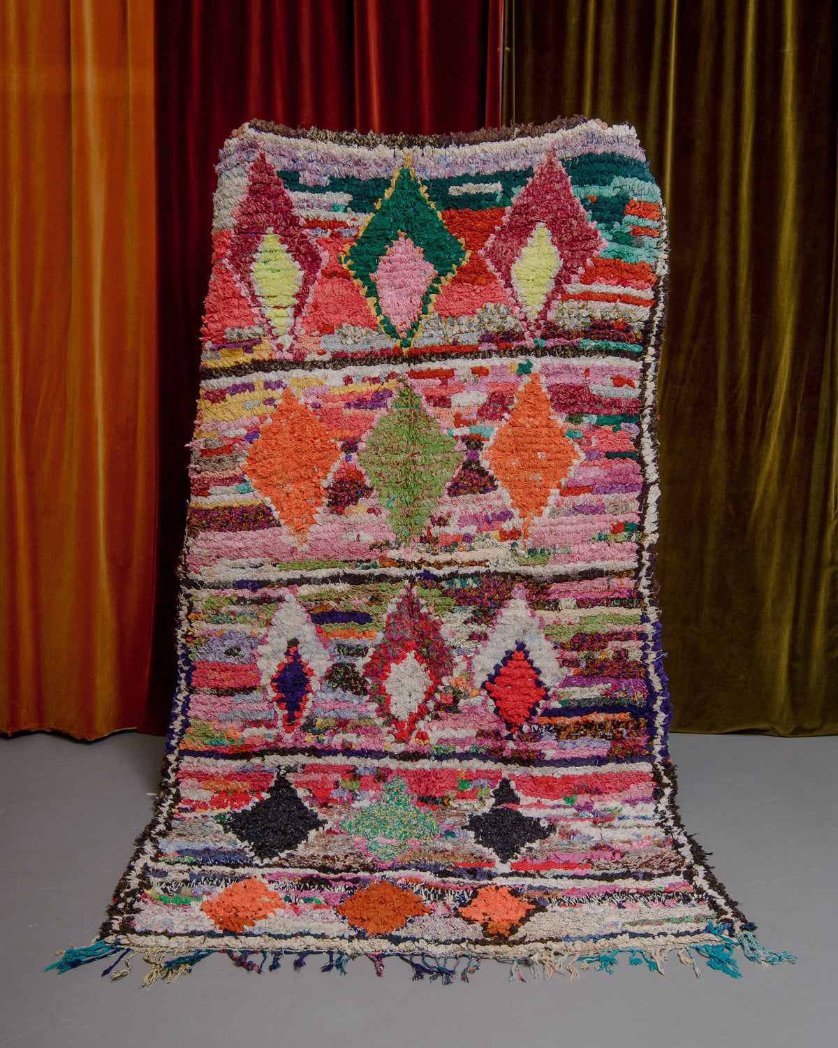 Boucherouite rug in varying shades of pink, orange and green, being held up in front of velvet curtains.