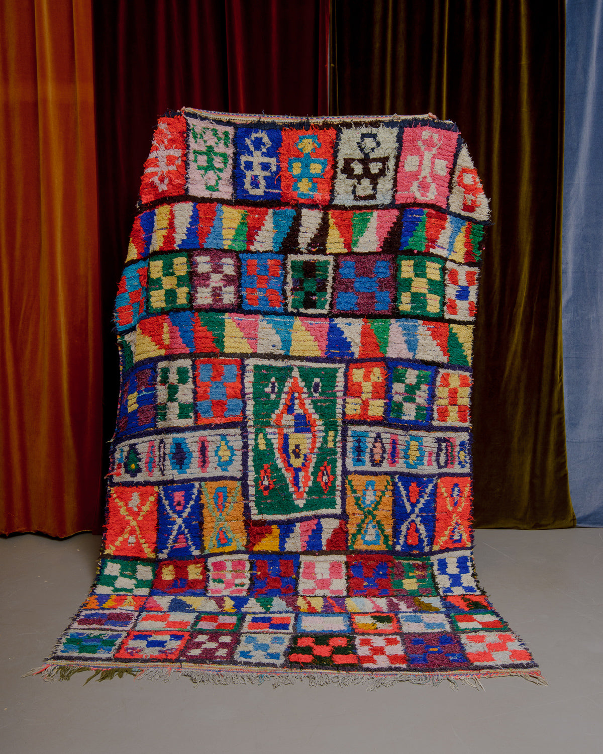 Boucherouite rug being held up in front of velvet curtains.