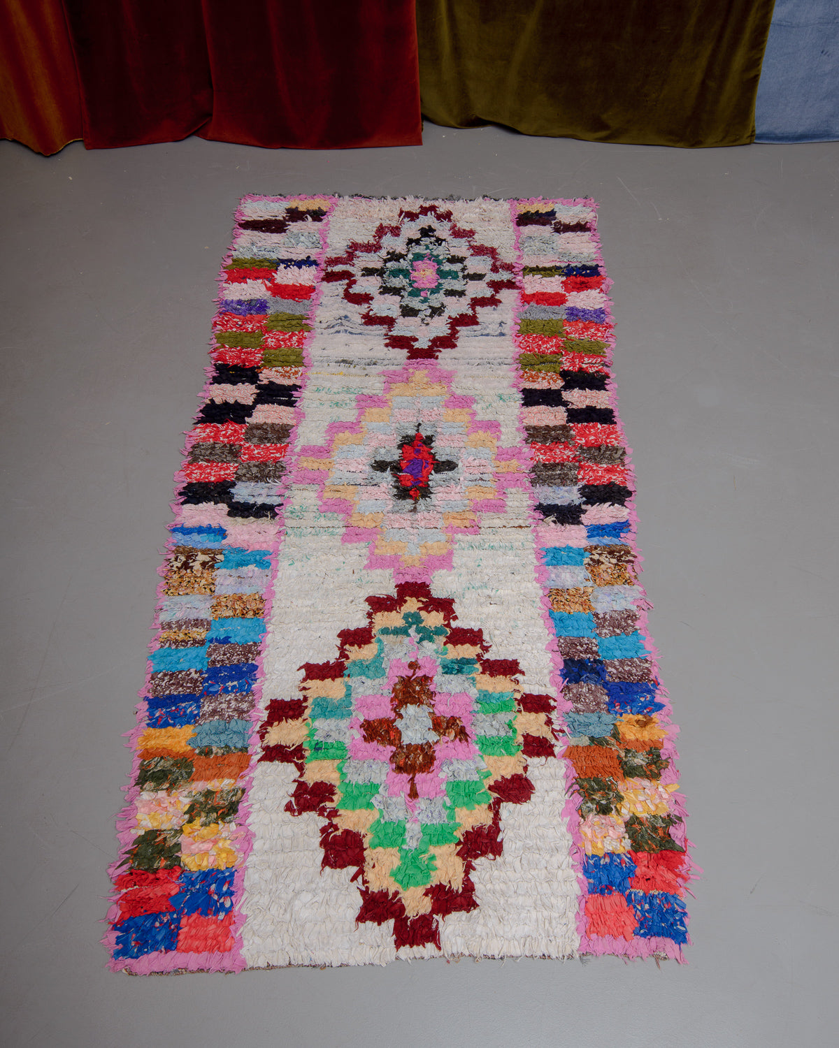 Vintage Berberlin boucherouite runner rug on grey floor in front of velvet curtain. 