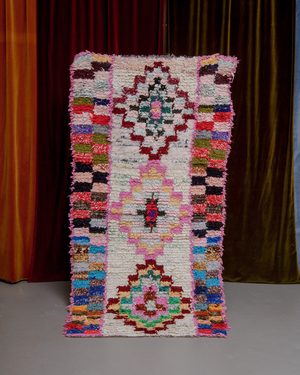 Vintage Berberlin boucherouite runner rug being held up in front of a velvet curtain. 
