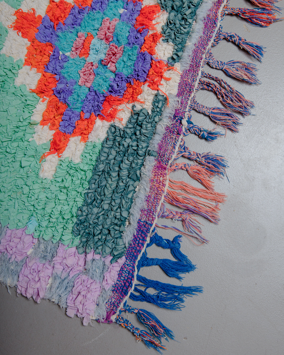 Corner of rug with lavender, turquoise, red, and blue with multicoloured tassels.
