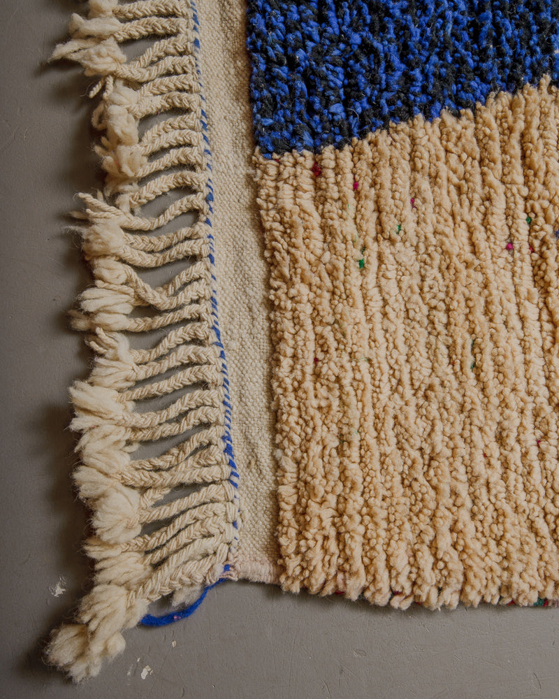 Corner of wool rug with cream and blue wool and dense tassels along the edge.
