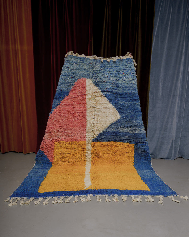 Berberlin Boujaad rug with bold forms in yellow, white and pink on a blue base being held in front of velvet curtains.