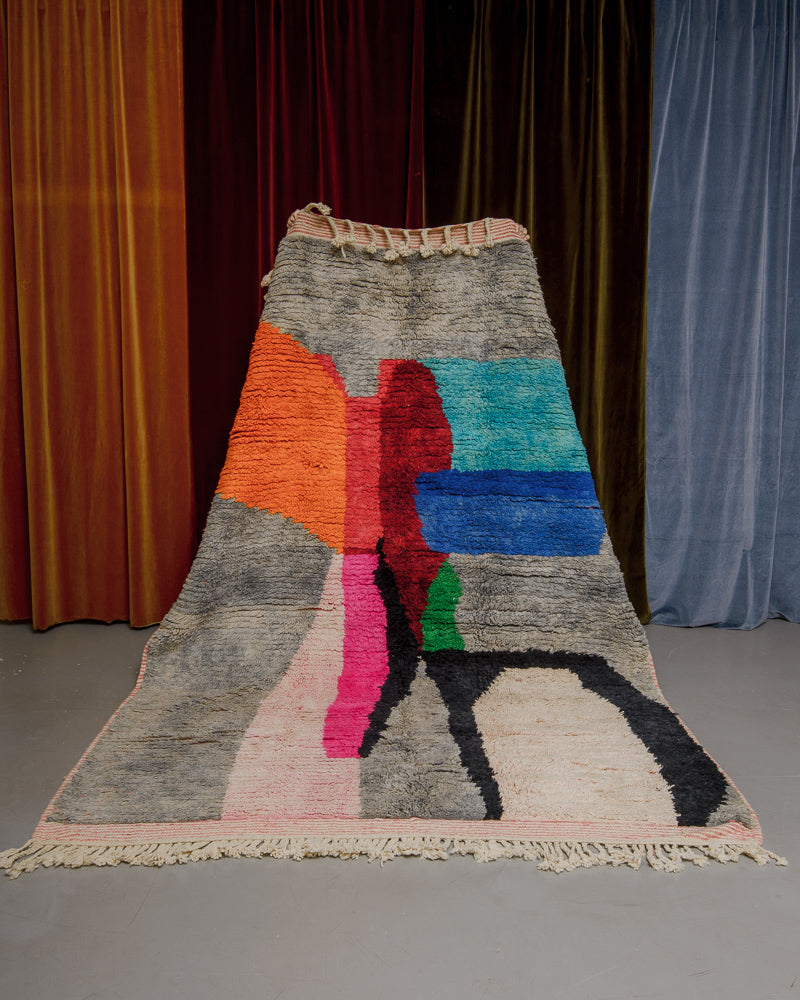 Berberlin Boujaad rug with bold forms in orange, red, blue, pink and grey being held in front of velvet curtains.