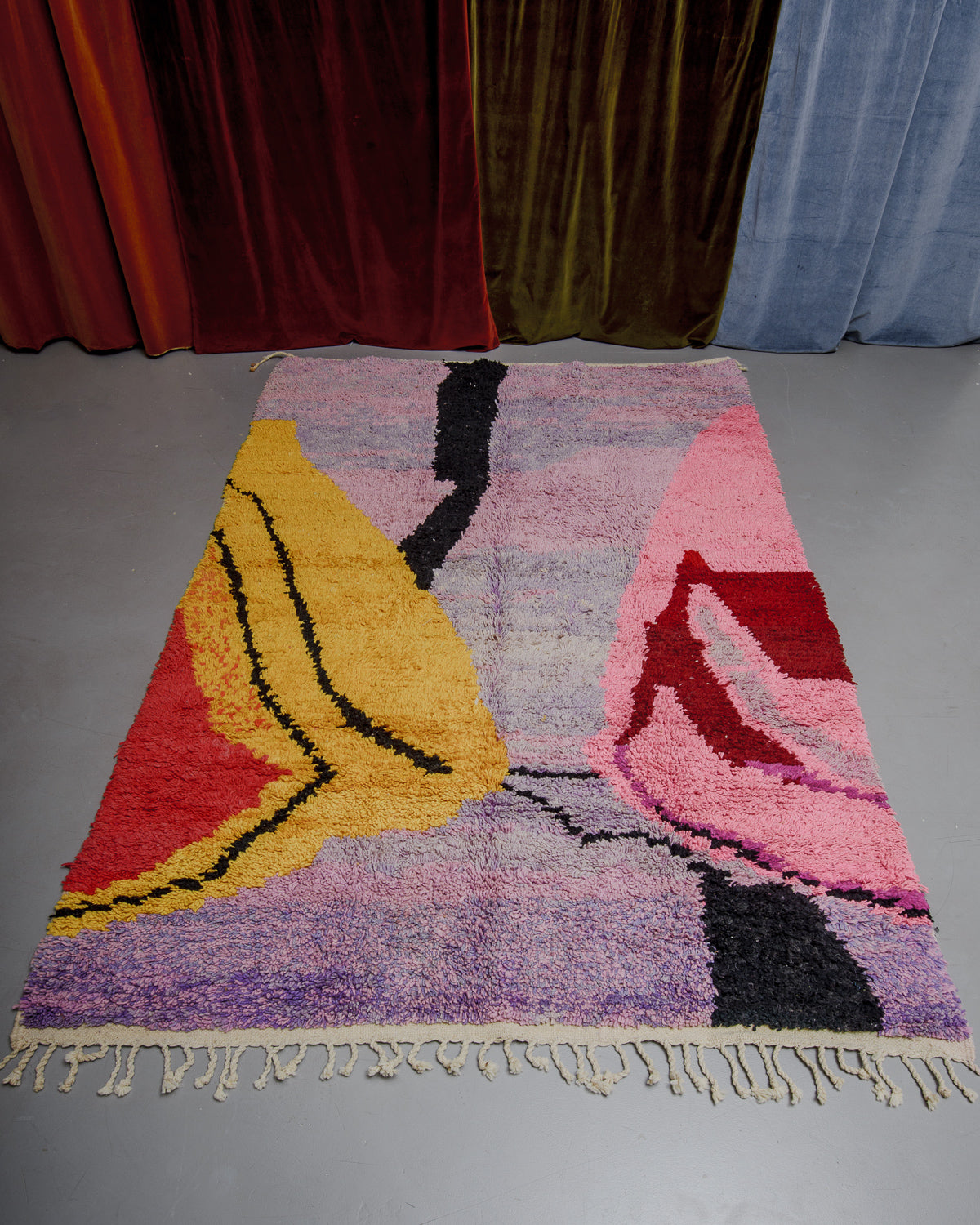 Berberlin Boujaad rug with bold, organic forms in lavender, pink, yellow and red on grey floor in front of velvet curtains.