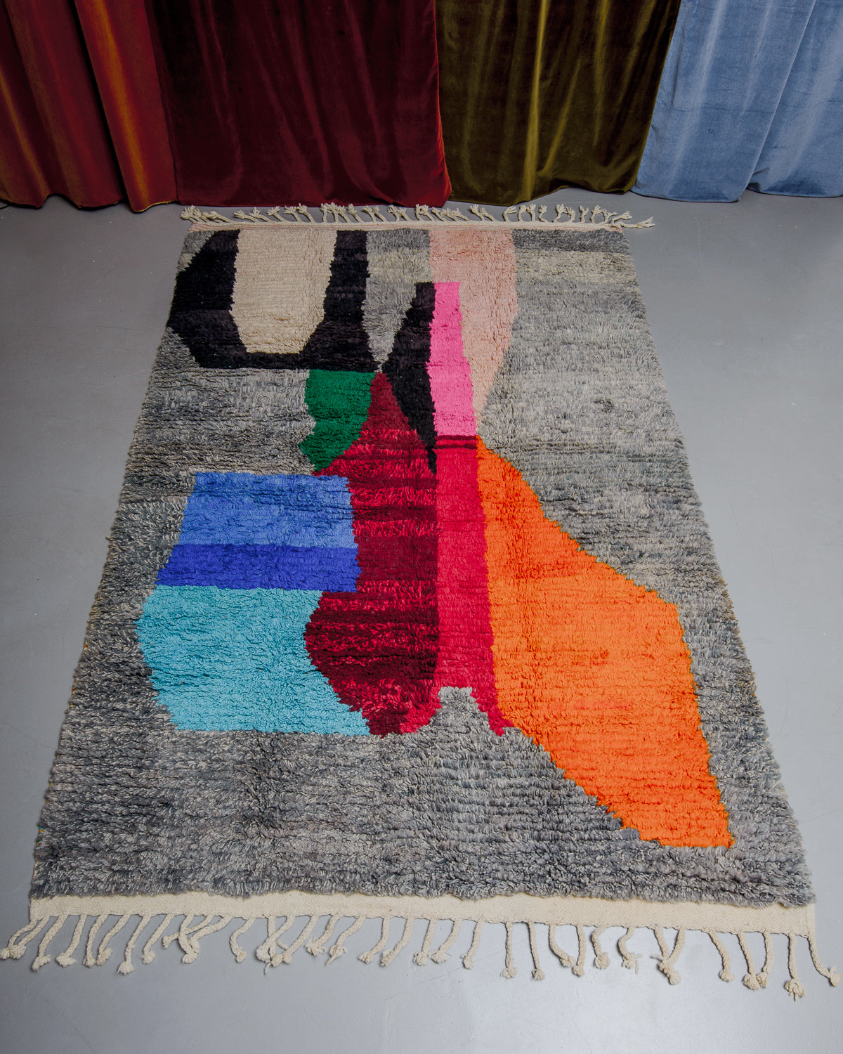 Berberlin Boujaad rug in grey with bold forms in orange, red, blue, pink and green on grey floor in front of velvet curtains.