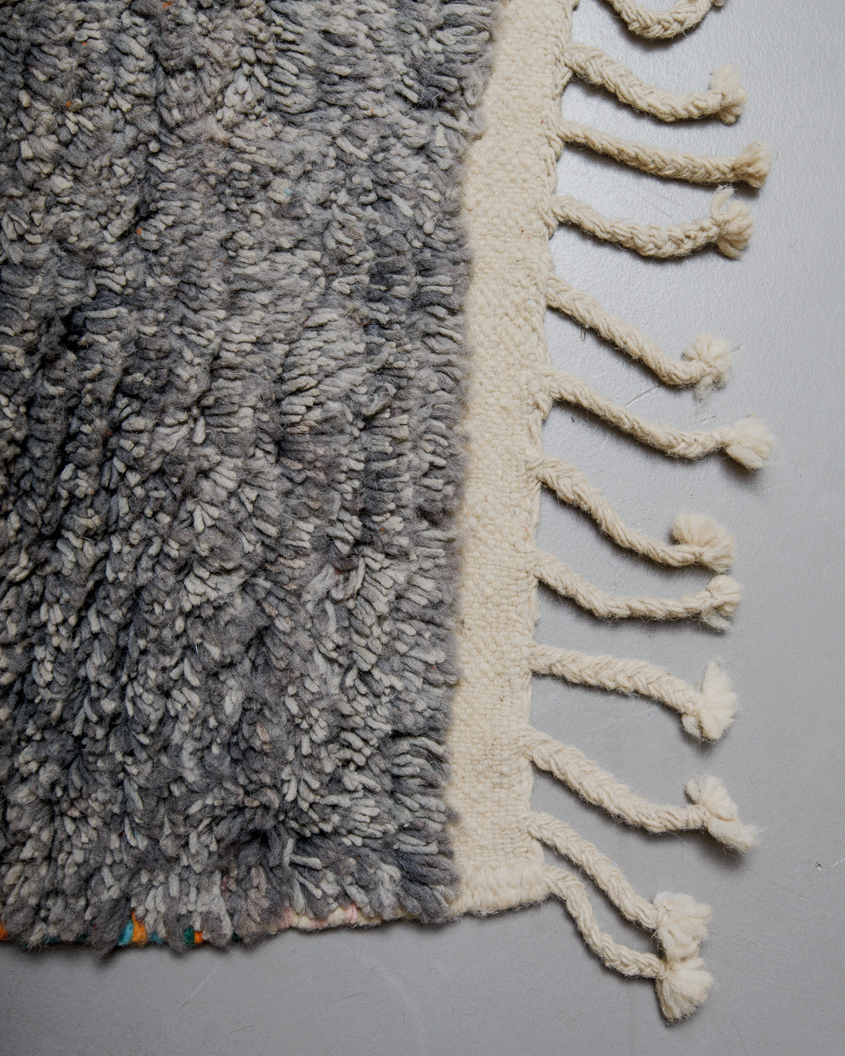 Corner of wool rug with grey pile and flat woven edge with natural wool tassels. 