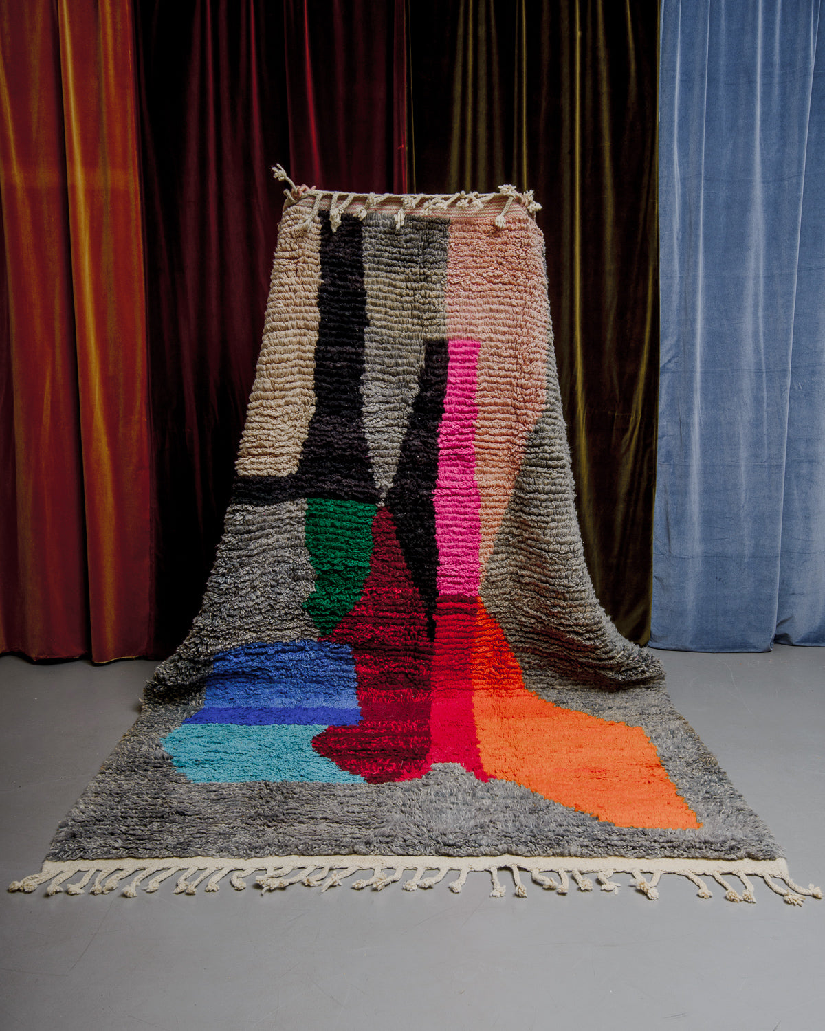 Berberlin Boujaad rug in grey with bold forms in orange, red, blue, pink and green being held in front of velvet curtains.