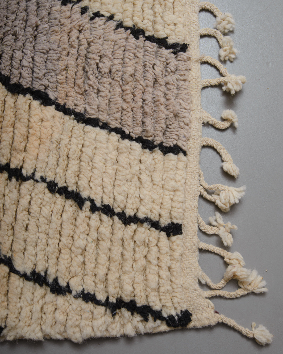 Corner of wool rug with cream and grey pile and flat woven edge with natural wool tassels. 