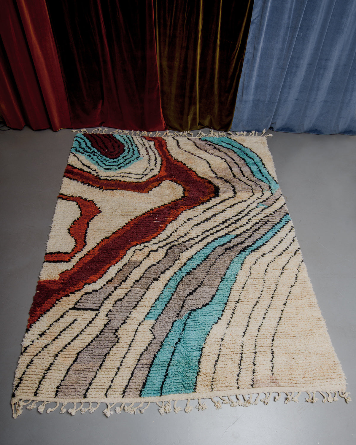 Berberlin Boujaad rug with bold, organic forms and lines in beige, grey, rust and turquoise on grey floor in front of velvet curtains.