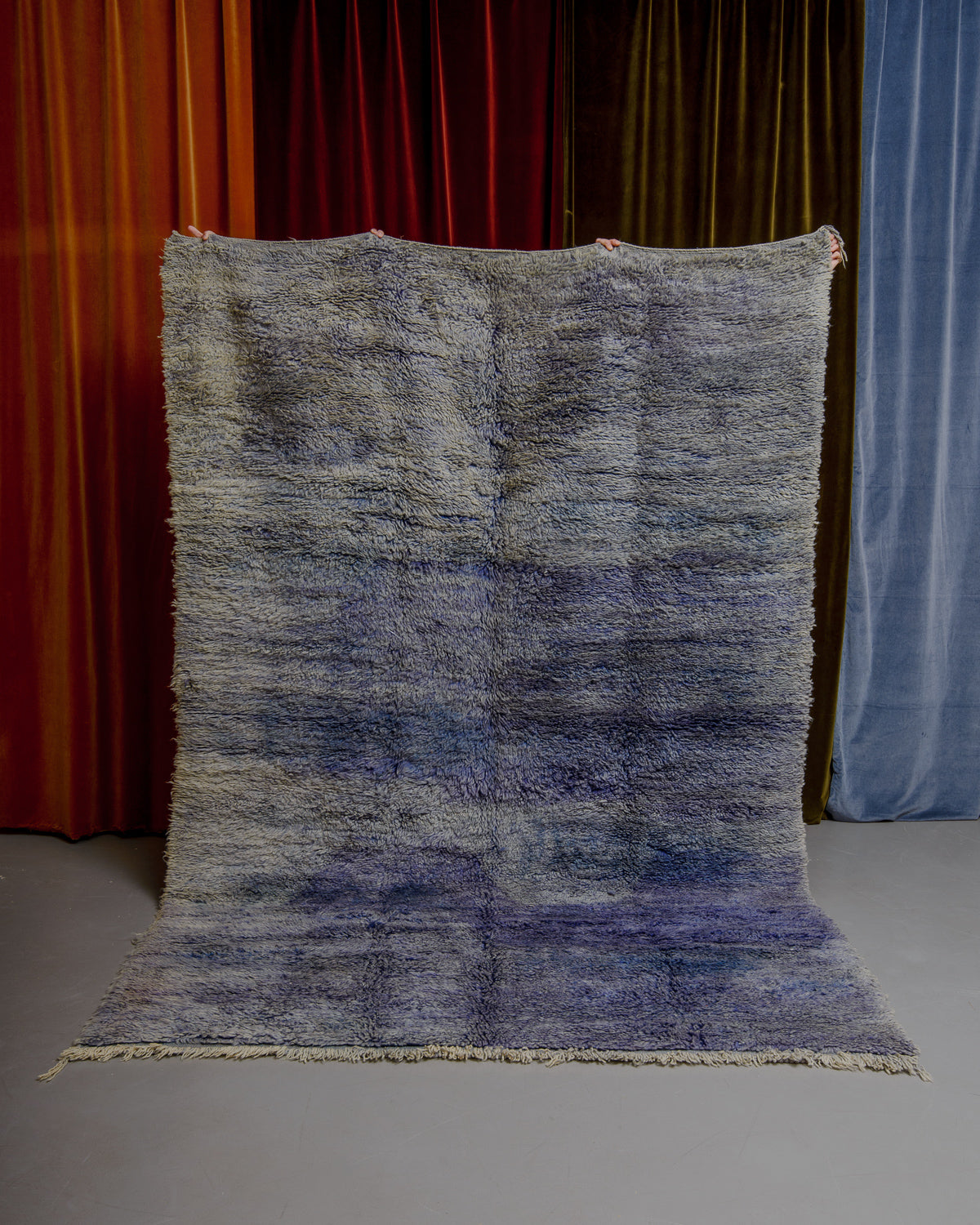 Berberlin Boujaad rug in marled blue and purple wool being held in front of velvet curtains.