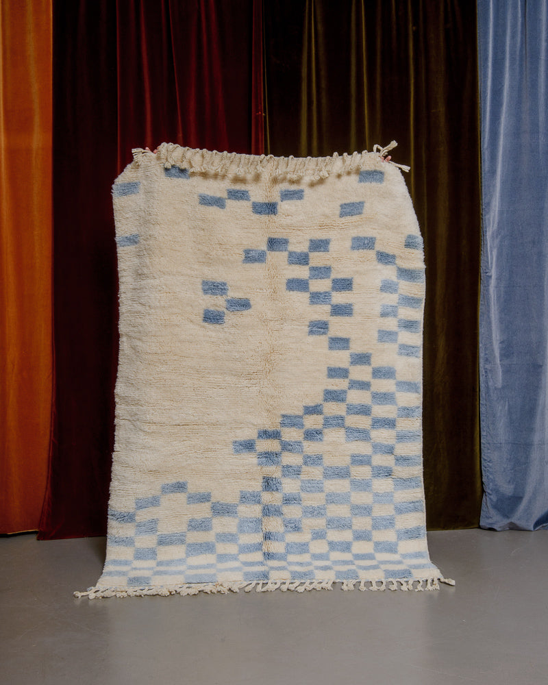 Cream wool rug with random blue checkered pattern in front of velvet curtains.