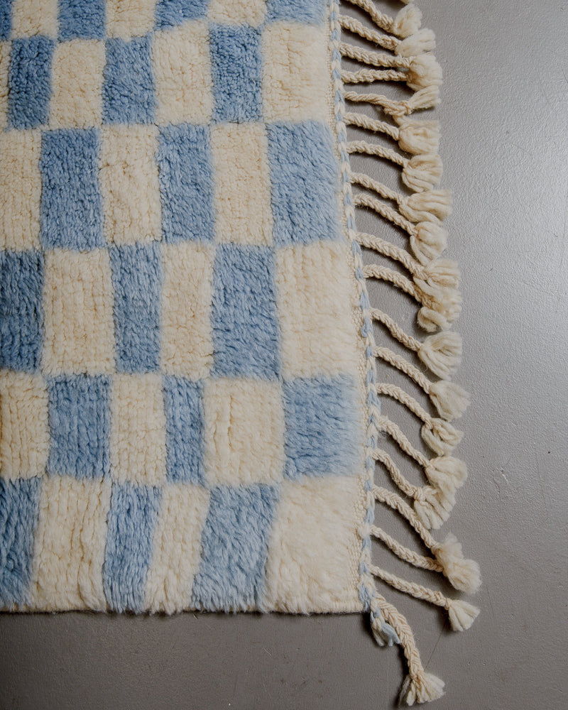 Corner of cream wool rug with sky blue pattern and natural wool tassels. 