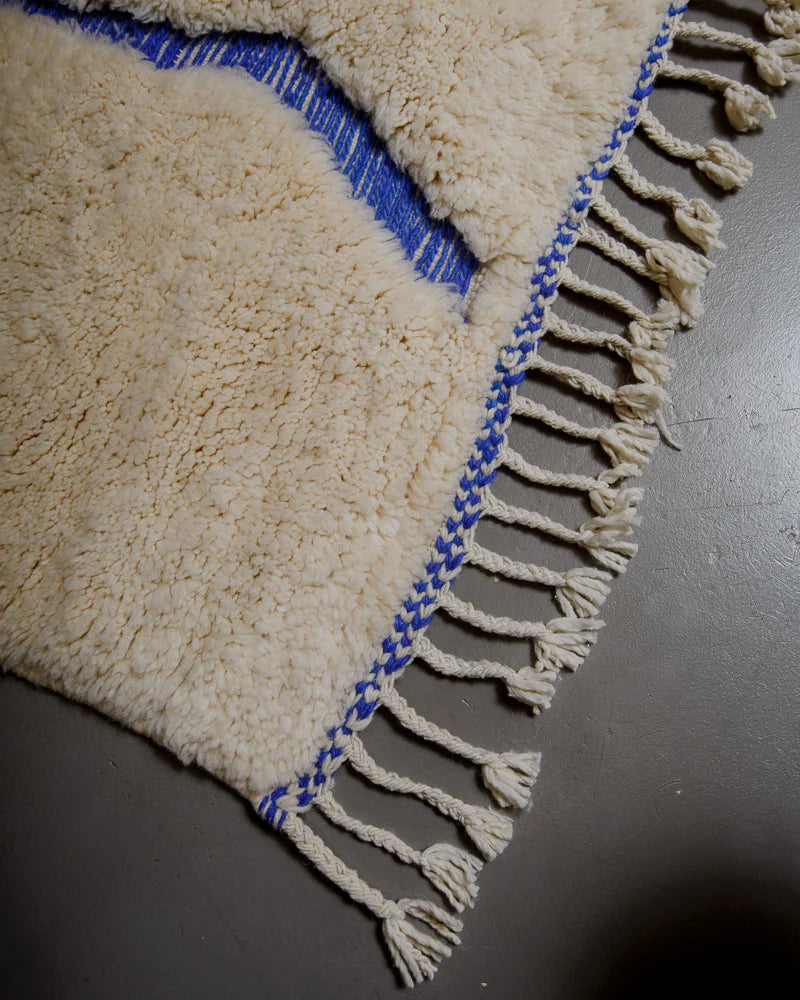 Corner of rug made of fluffy cream wool pile and woven blue lines with natural wool tassels along the edge.