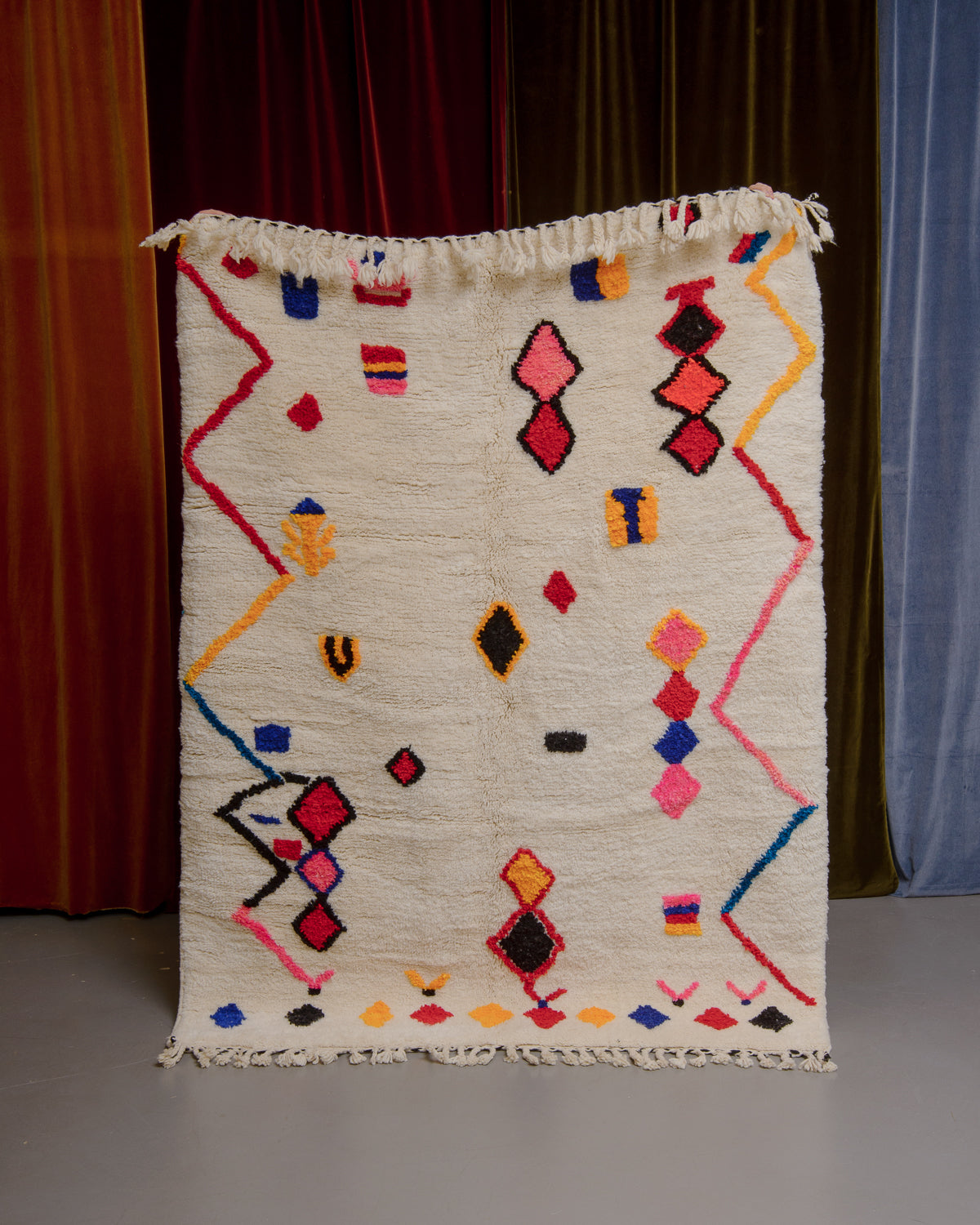 Berberlin Finest rug with a heavy cream pile and colourful design in cotton being held in front of velvet curtains. 