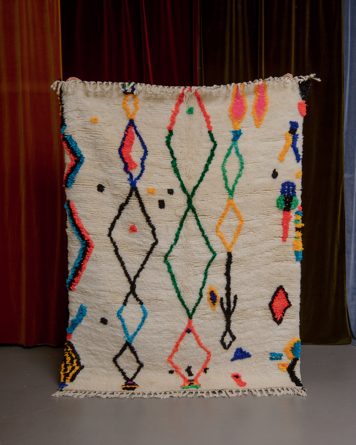 Berberlin Finest rug with a heavy cream pile and colourful design in cotton being held in front of velvet curtains. 