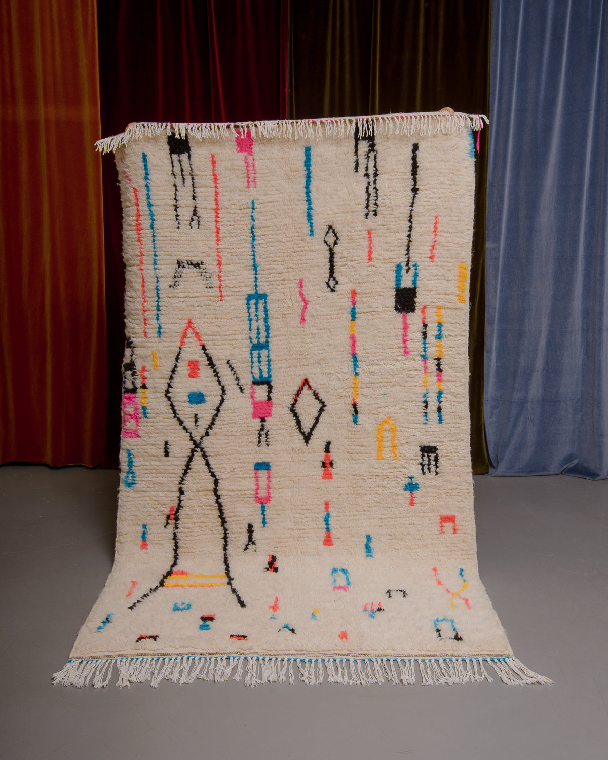 Azilal rug with colourful, abstract design being held up in front of a wall of velvet curtains.