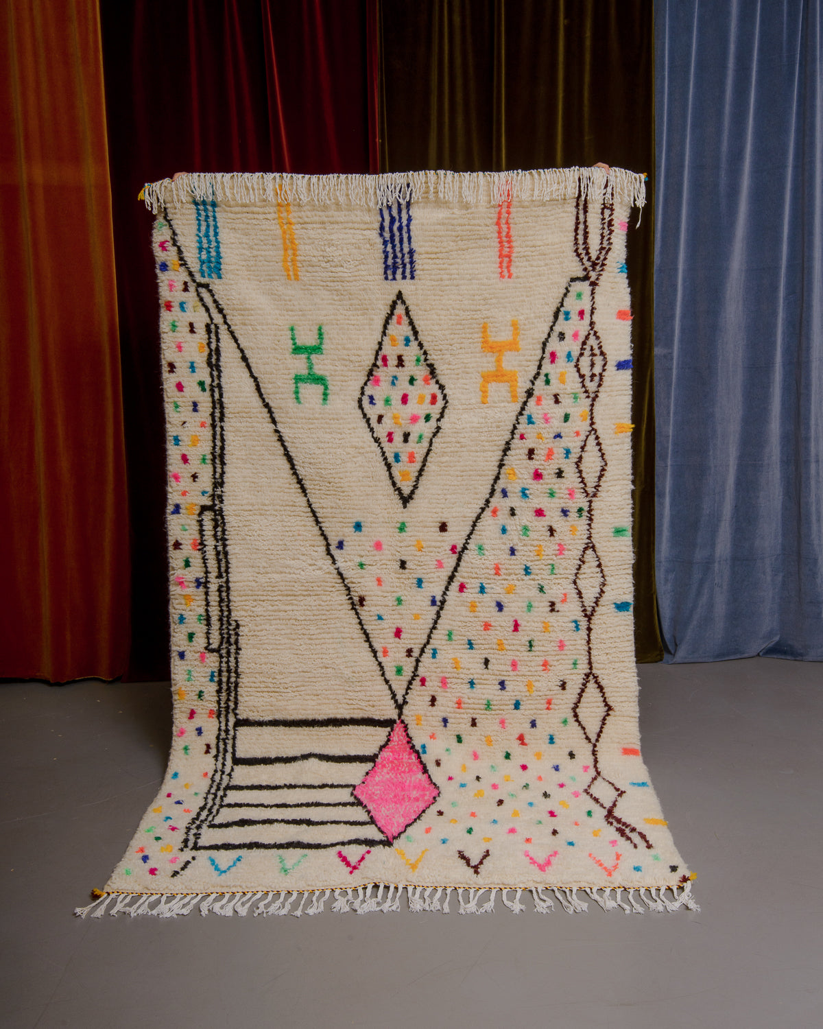 Azilal rug with colourful, abstract design being held up in front of a wall of velvet curtains.