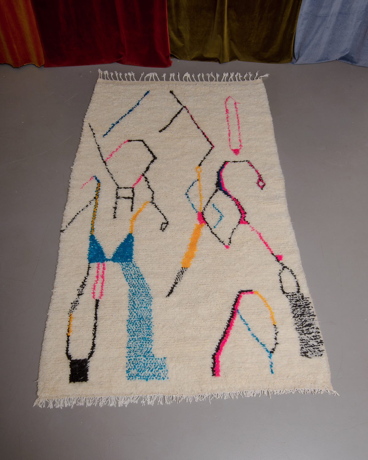Azilal rug with colourful, abstract design laid flat on grey floor in front of a wall of velvet curtains.