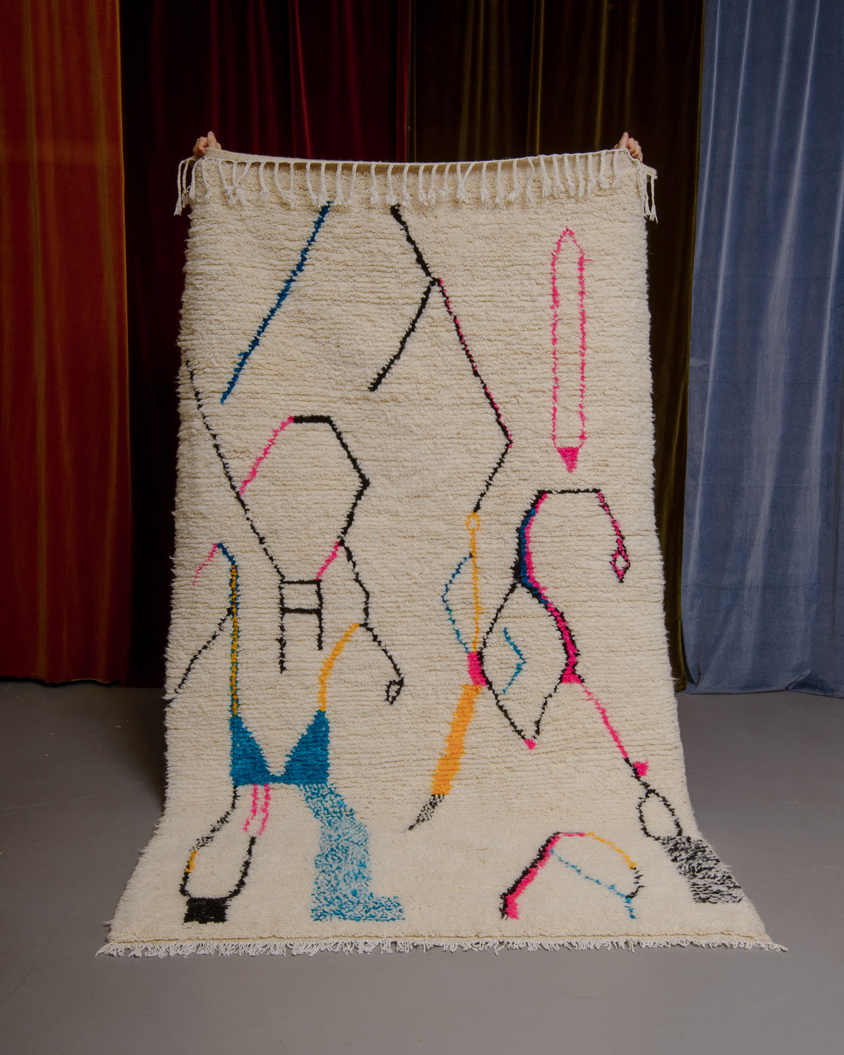 Azilal rug with colourful, abstract design being held up in front of a wall of velvet curtains.