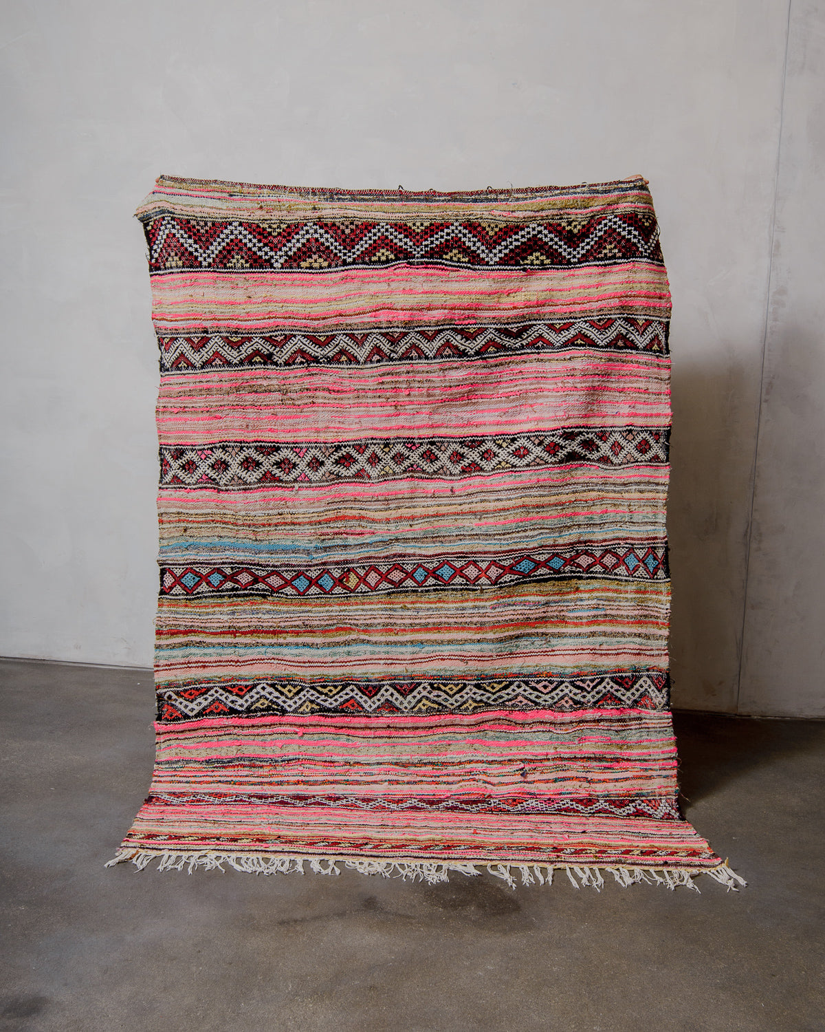 Multicoloured striped rug with traditional patterns and cotton fringe on a grey floor against a white wall.