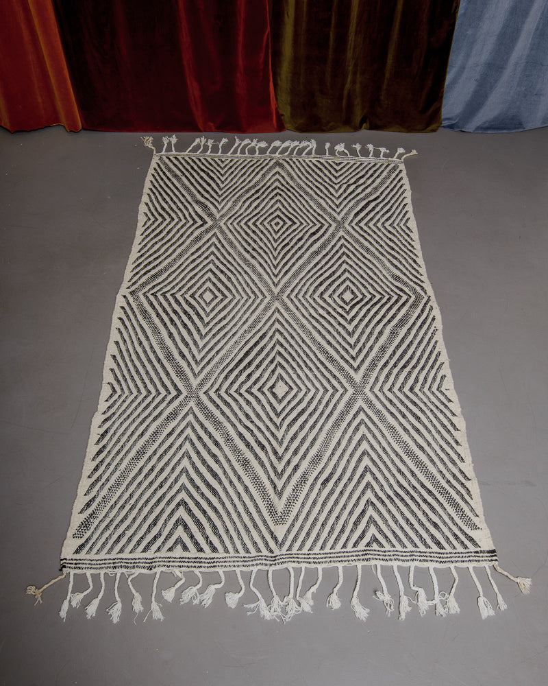 Black and white Kelim rug on grey floor in front of velvet curtains. 