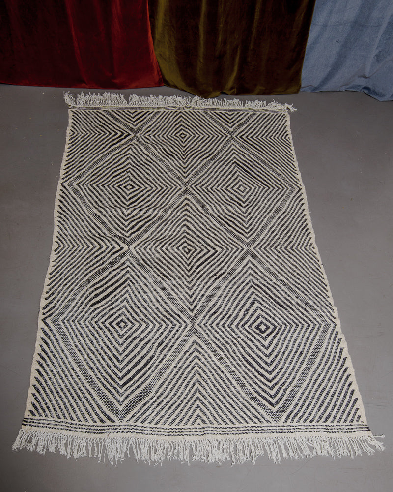 Black and white Kelim rug on grey floor in front of velvet curtains. 