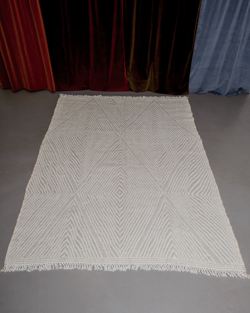 Grey and white Kelim rug on grey floor in front of velvet curtains. 