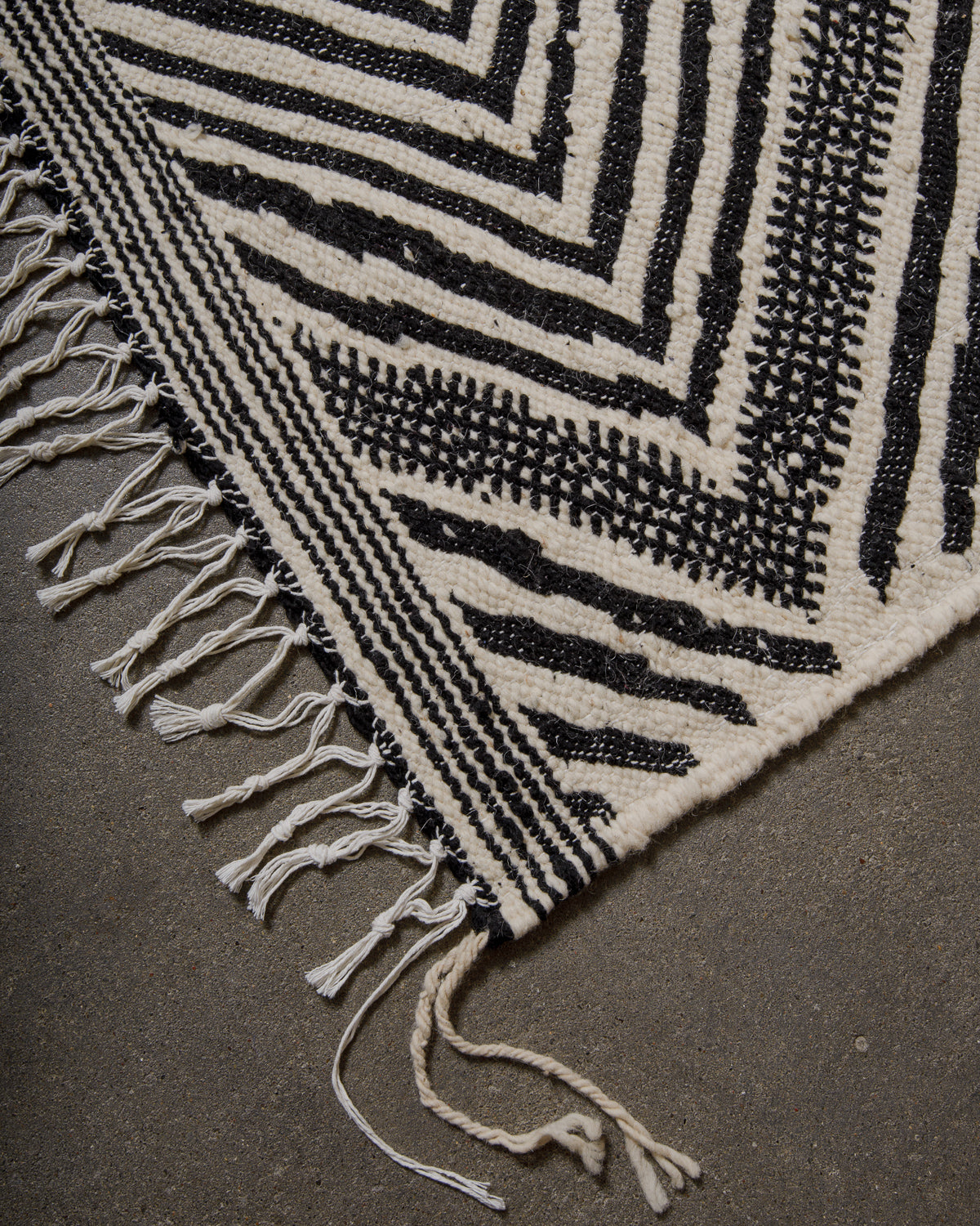Black and white patterned Kelim rug with fringe on a dark surface.