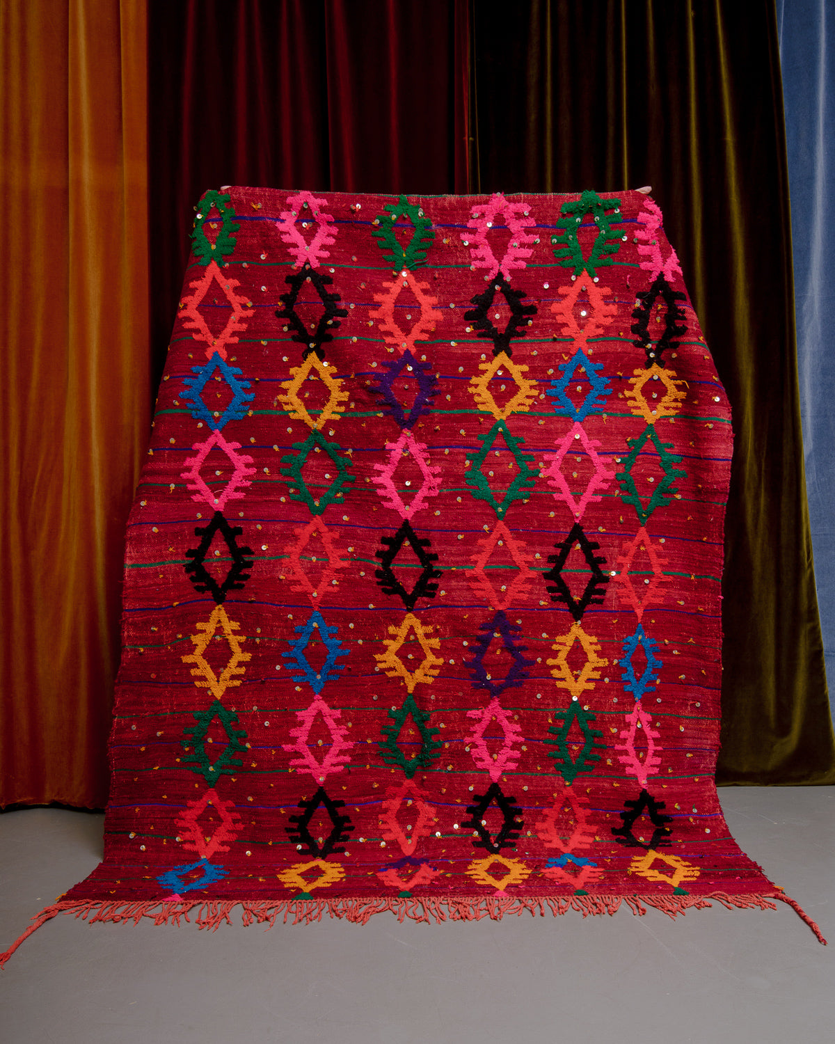 Vintage Kelim rug in bright red, green, orange, pink and blue being held in front of velvet curtains. 
