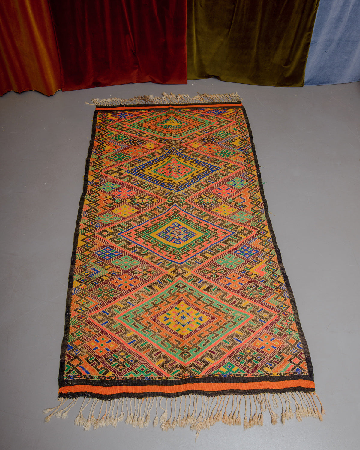 Vintage Kelim rug in bright yellow, orange, blue and green on grey floor with velvet curtains in the background. 