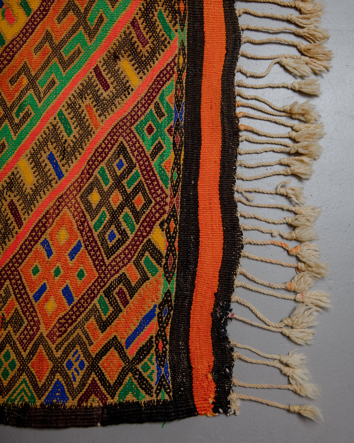 Corner of Vintage Kelim rug in bright yellow, orange, blue and green with a black border and cream wool tassels.